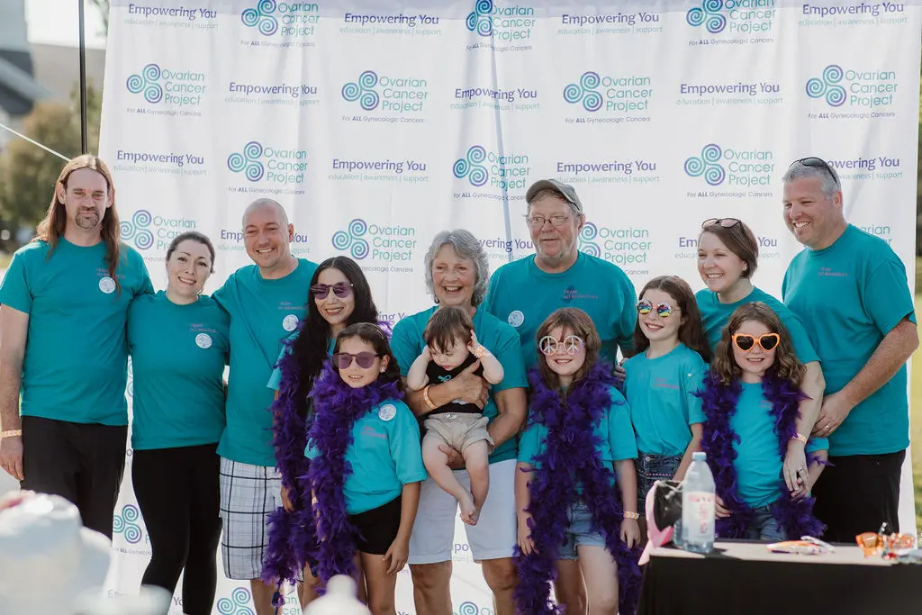 Group of people in teal shirts smiling at an ovarian cancer awareness event. Some wear purple boas. Background has supportive messaging.